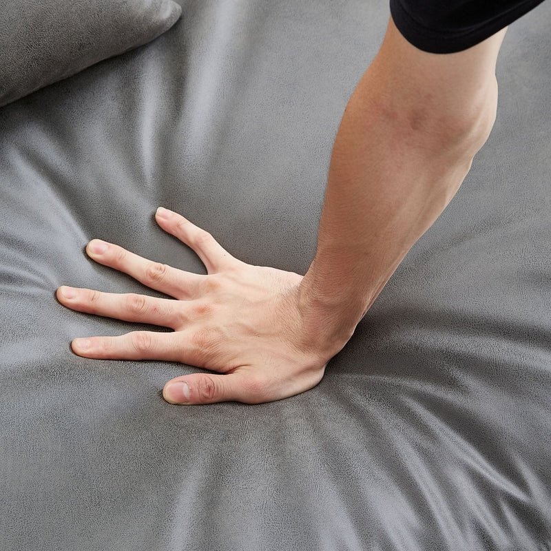 A hand presses down on **The 5th Open 116" Left Facing Sectional** in a soft gray material, creating visible indents. The surface appears smooth and cushioned, suggesting comfort and support.