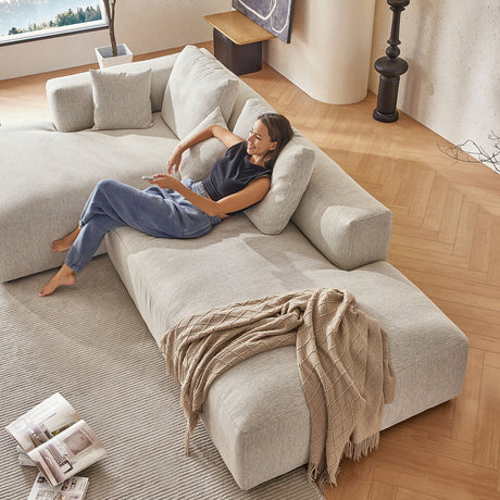 A woman in casual clothing relaxes on "The 5th Open 116" Left Facing Sectional," a large, light gray sofa in a modern living room. A beige throw is draped over the sofa, while magazines lie open on a nearby rug, and soft natural light fills the space.