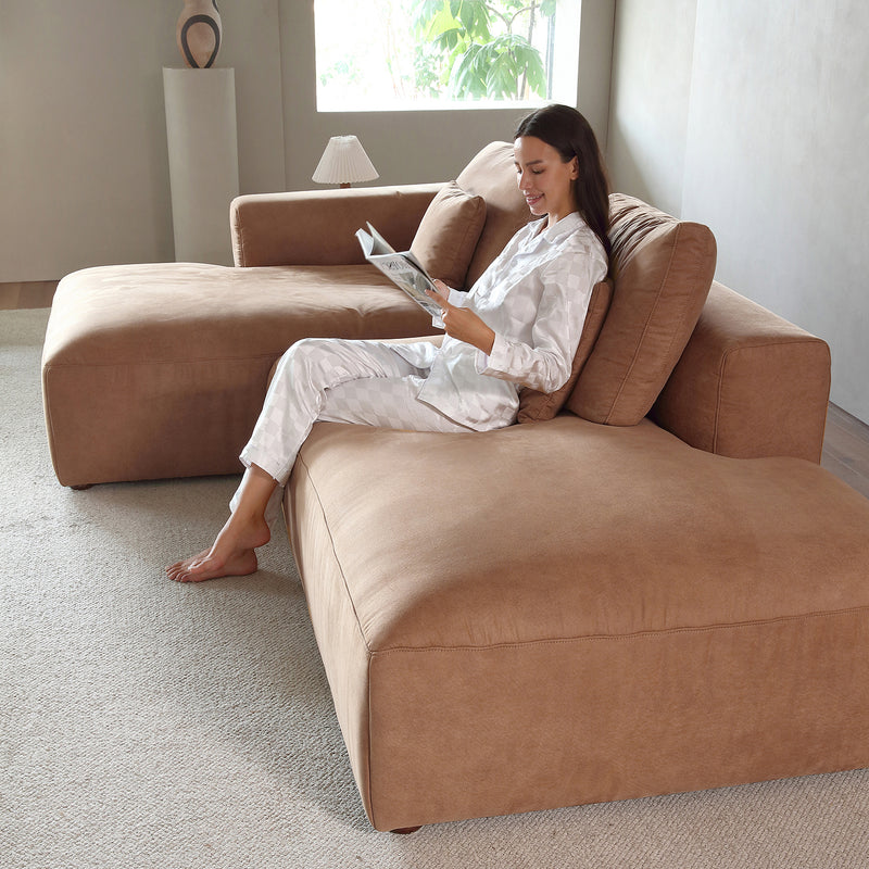Dressed in light pajamas, an individual relaxes on The 5th Open 116" Left Facing Sectional, a spacious L-shaped sofa in brown, while enjoying a magazine. The room features neutral tones with soft lighting and minimalistic decor, including a small lamp and decorative objects on a shelf. A window allows natural light to filter in.
