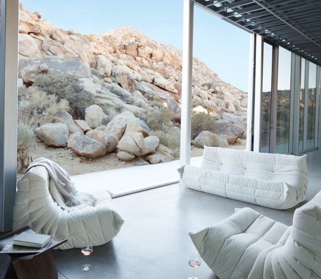 A modern living room featuring the Togo Sofa in Opera Leather and large glass windows offers a view of a rocky desert landscape. Sunlight brightens the space, accentuating the sleek white flooring and minimalist decor, with a couple of glasses resting on the floor.