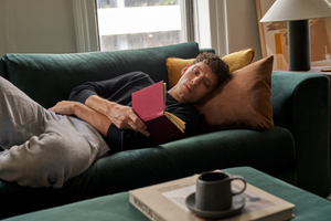 A person with curly hair is lying on The Elevate Sofa, holding an open book with a red cover. They are dressed in a black sweatshirt and gray sweatpants. On the nearby coffee table, there's a mug and another book. The room is softly illuminated by natural light.