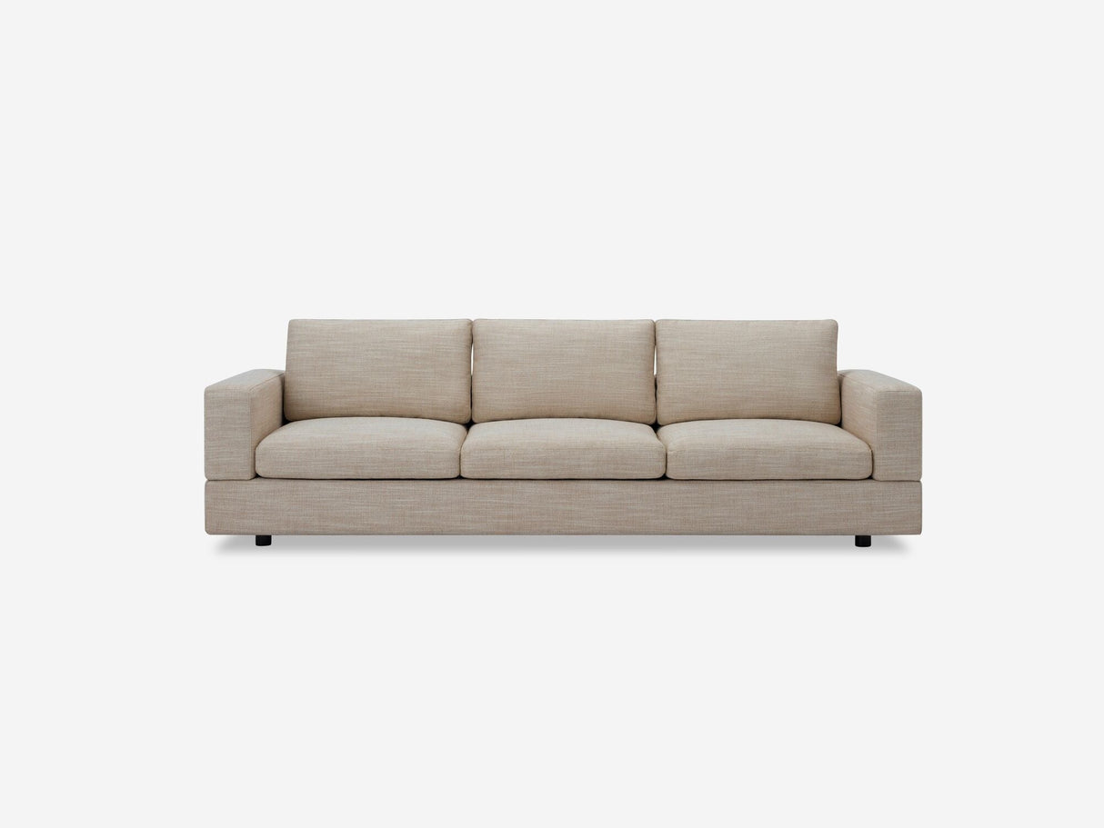 A Stage Track Sofa, featuring a simple and modern design with a beige color, three seats, rectangular cushions, and padded armrests on each side, is set against a plain white background.