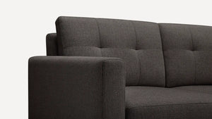 A close-up view of the Nomad Fabric Sofa in dark gray, featuring a tufted backrest and a thick armrest, seen from the left side against a plain white background.