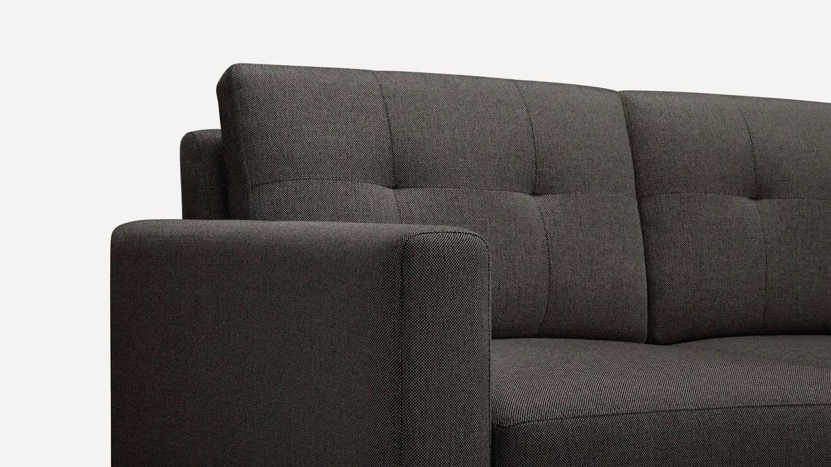 A close-up view of the Nomad Fabric Sofa in dark gray, featuring a tufted backrest and a thick armrest, seen from the left side against a plain white background.
