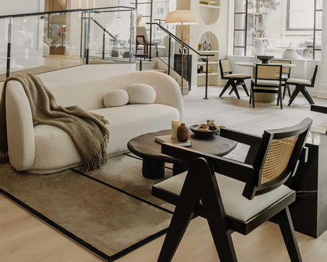 A sophisticated living room features a cream-colored Diane Sofa with a dark wood coffee table and modern black chairs. A cozy throw is draped over the sofa. Large windows illuminate the space, revealing a dining area in the background.