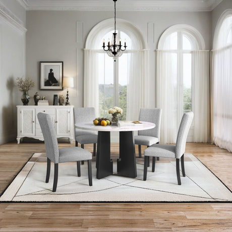 A modern dining room features the 5 Piece Round Dining Table Set with a white table and four gray upholstered chairs, all on a light geometric-patterned rug. A chandelier hangs above, while a sideboard adorned with art and a lamp rests against the wall with large arched windows.