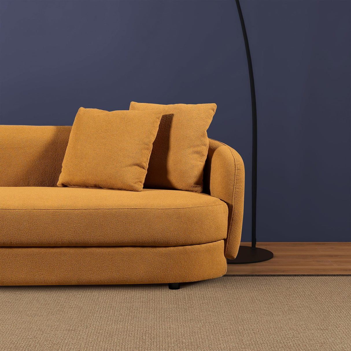 A mustard-yellow Chandler Sectional Sofa with two matching cushions sits on a beige carpet against a dark blue wall, accompanied by a sleek black floor lamp with a curved stand behind it.