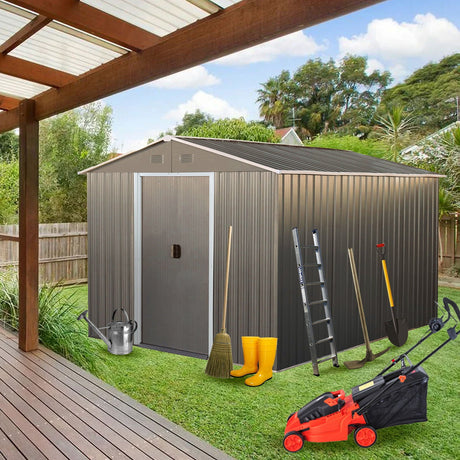 An 8X10Ft Outdoor Metal Storage Shed in gray is near a wooden deck. In front, gardening tools like a broom, shovel, rake, ladder, watering can, and lawnmower are visible with yellow boots beside them. Trees and a fence provide the backdrop.