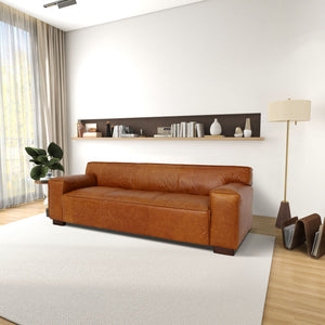 A spacious living room features the Morgan - Waxy Sofa in Cognac centered on a white rug. A floor lamp stands beside it, with books and decor on a wooden shelf above. Sunlight filters through a large window with beige curtains, while a plant adds greenery to the space.