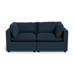 The Kova Pillow Cushion Sofa 86" is a modern, dark blue upholstered sofa with wide armrests and four large cushions, displayed front-facing against a plain black background.
