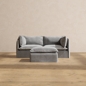 The Modular Performance 2-Seater in minimalist gray, complete with matching pillows, is centered against a beige wall. The set includes a small gray ottoman positioned in front of the sofa. This arrangement sits atop light wooden flooring, creating a simple and modern aesthetic.