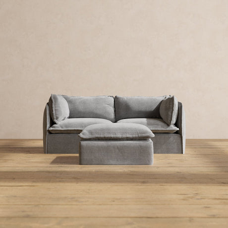 A chic gray Modular Performance 2-Seater with coordinated cushions is set against a beige backdrop. In front of the sofa, a square gray ottoman complements the scene, resting on a light wooden floor. The arrangement exudes a minimalist and tranquil ambiance.