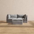 The Modular Performance 2-Seater in minimalist gray, complete with matching pillows, is centered against a beige wall. The set includes a small gray ottoman positioned in front of the sofa. This arrangement sits atop light wooden flooring, creating a simple and modern aesthetic.