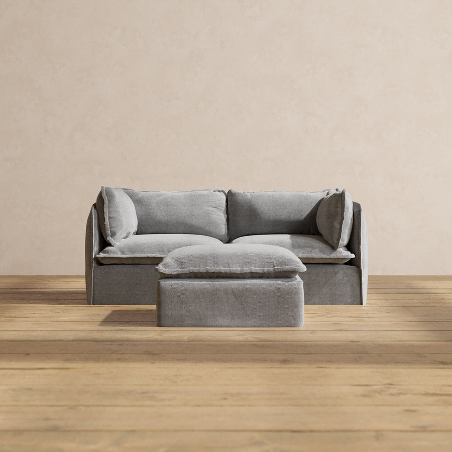 The Modular Performance 2-Seater in minimalist gray, complete with matching pillows, is centered against a beige wall. The set includes a small gray ottoman positioned in front of the sofa. This arrangement sits atop light wooden flooring, creating a simple and modern aesthetic.