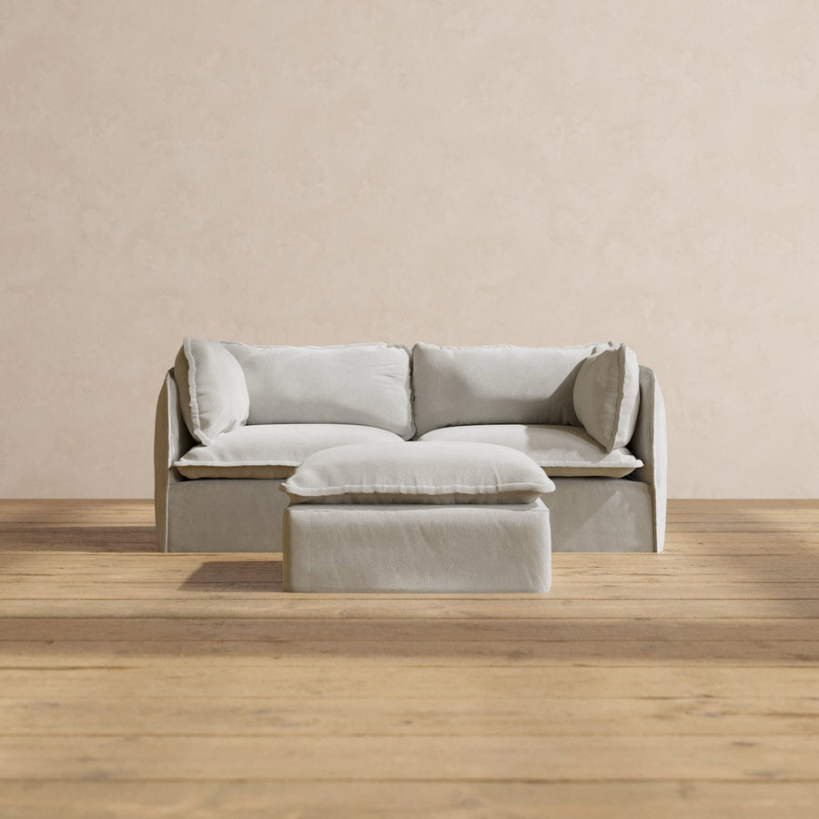 A Modular Performance 2-Seater in a minimalist cream color, complemented by matching cushions, is situated against a pale beige wall, with the accompanying square ottoman placed in front of it. The floor features light wood planks.