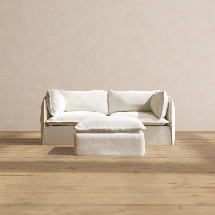 A Modular Performance 2-Seater in a minimalist beige design with matching cushions is set on a wooden floor against a plain beige wall. In front of the sofa sits the matching rectangular ottoman with a soft cushion that enhances the neutral color scheme.