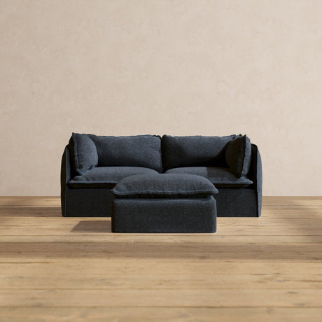 A Modular Performance 2-Seater in dark blue, complete with matching cushions, is positioned on a wooden floor against a beige wall. A small square ottoman in the same color is placed in front of the sofa.