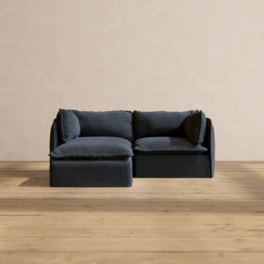 The Modular Performance 2-Seater Sectional, in a contemporary dark grey with plush cushions, rests on a light wood floor beside a plain beige wall, crafting a minimalist and cozy atmosphere.