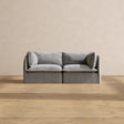 A Modular Performance 2-Seater with Sink-in Comfort Cushion in a modern gray design rests on a wooden floor. The plain beige wall in the background enhances the minimalist and cozy ambiance.