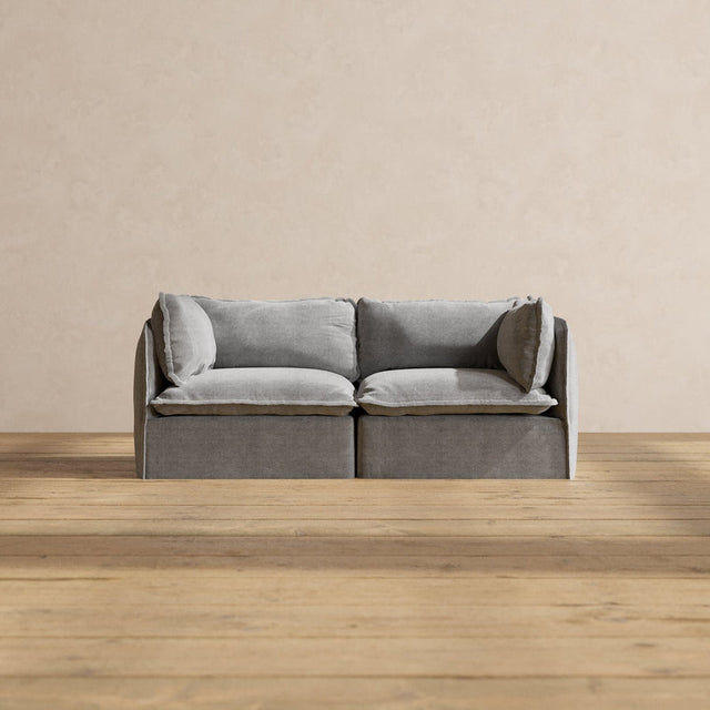 A Modular Performance 2-Seater with Sink-in Comfort Cushion in a modern gray design rests on a wooden floor. The plain beige wall in the background enhances the minimalist and cozy ambiance.