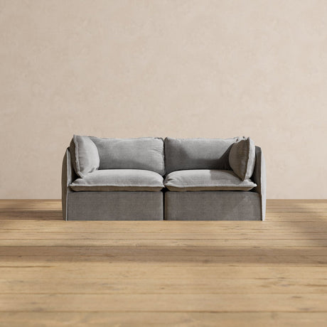 A Modular Performance 2-Seater with Sink-in Comfort Cushion, featuring a modern gray design and plush cushions, sits on a wooden floor against a beige wall. The minimalist design offers a cozy and stylish seating option.
