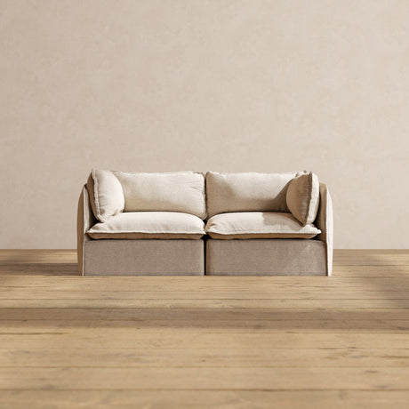 A Modular Performance 2-Seater with Sink-in Comfort Cushion, featuring a modern and minimalist design in beige, is positioned on a wooden floor against a plain light-colored wall. It includes two large cushions and an armrest on each side.