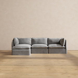 A sleek Modular Performance 3-Seater Sectional in a minimalist gray design is positioned on a wooden floor against a neutral, light beige wall. This sectional includes three seating sections with a chaise lounge on the left side.