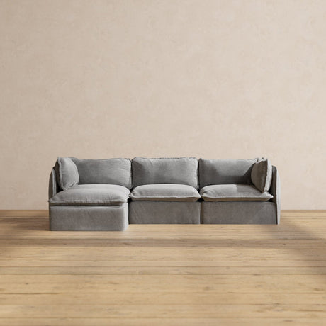 A sleek Modular Performance 3-Seater Sectional in a minimalist gray design is positioned on a wooden floor against a neutral, light beige wall. This sectional includes three seating sections with a chaise lounge on the left side.