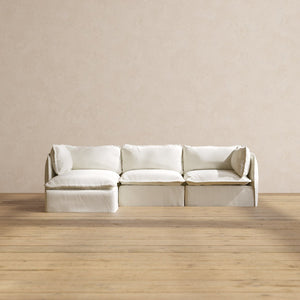 In a minimalist room, a Modular Performance 3-Seater Sectional is positioned against a beige wall on a wooden floor. The sectional boasts plush cushions and an inviting appearance.