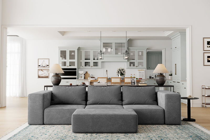 A modern living room showcases the Atlas Modular Sofa by Acanva, a large gray sectional facing the viewer, complemented by a light blue patterned rug beneath it. In the background, an open doorway reveals a spacious kitchen with light gray cabinetry and pendant lights.