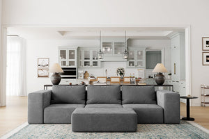 A modern living room showcases the Atlas Modular Sofa by Acanva, a large gray sectional facing the viewer, complemented by a light blue patterned rug beneath it. In the background, an open doorway reveals a spacious kitchen with light gray cabinetry and pendant lights.