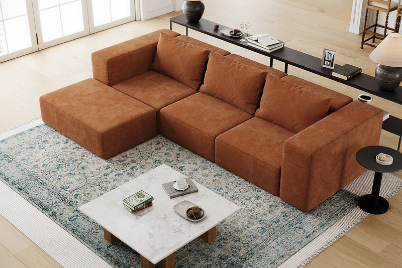 A top view of a cozy living room features the Atlas Modular Sofa by Acanva in a large brown sectional style, placed on a blue patterned area rug. In front of the sofa sits a marble coffee table adorned with books and a candle, while along the back wall near wide windows is a console table decorated with various items.