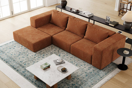 A top view of a cozy living room features the Atlas Modular Sofa by Acanva in a large brown sectional style, placed on a blue patterned area rug. In front of the sofa sits a marble coffee table adorned with books and a candle, while along the back wall near wide windows is a console table decorated with various items.