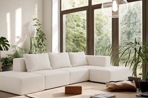 A cozy living room featuring an Atlas Modular Sofa by Acanva in a large, white configuration, complemented by green plants and a wooden coffee table. Natural light floods through floor-to-ceiling windows, accentuating the neutral decor and soft rug.
