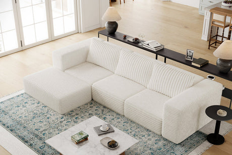 A cozy living room features the Atlas Modular Sofa by Acanva, with a cream-colored ribbed design, placed atop a patterned blue rug. A marble coffee table nearby holds books, snacks, and a cup. Behind the sofa stands a console table adorned with lamps and decor items. The space is filled with natural light streaming in through large windows.