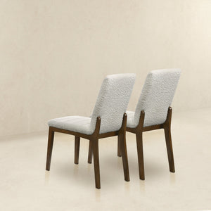 The Ines Modern Dining Chair set of 2 features light textured upholstery and dark wooden legs, positioned on a polished light floor against a plain beige wall. The chairs are viewed from the back, slightly angled to the left.