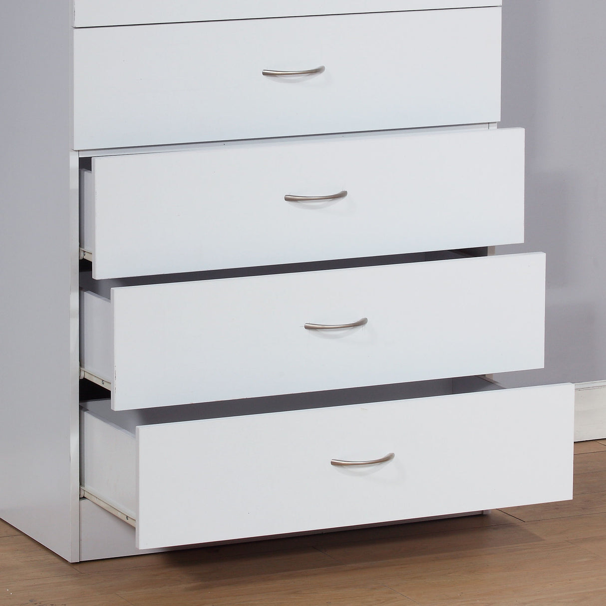 A white 5-drawer chest with silver metal handles rests against a light wall. Three drawers are open, one is closed, and the top drawer locks. The floor features light wood.