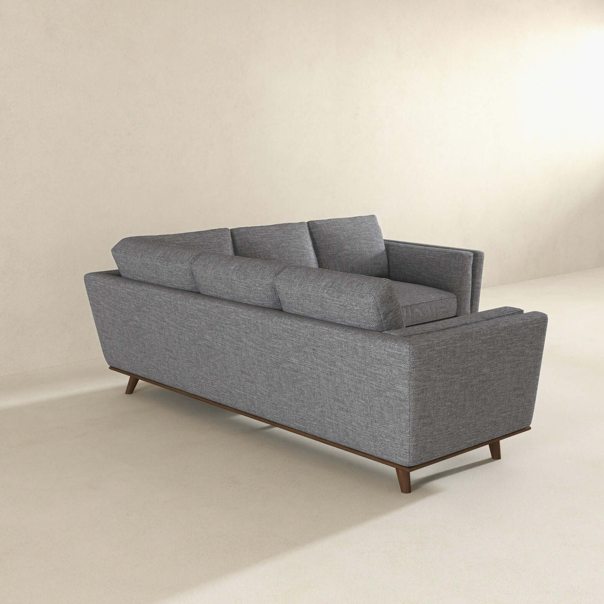 The Erman - Pillow Back Corner Sofa in dark gray, featuring three back cushions and tapered wooden legs, sits against a light wall. Its minimalist design complements the spacious, neutral-toned room.