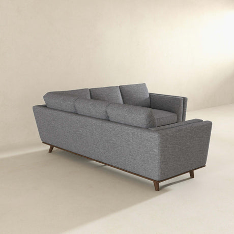 The Erman - Pillow Back Corner Sofa in dark gray, featuring three back cushions and tapered wooden legs, sits against a light wall. Its minimalist design complements the spacious, neutral-toned room.