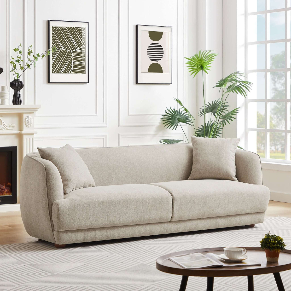 The Larisa Mid-Century Modern Linen Sofa, complemented by cushions, enhances a contemporary living room adorned with two abstract framed artworks and a fireplace with a floral vase. Nearby, a large window illuminates the round coffee table in the foreground stacked with books.