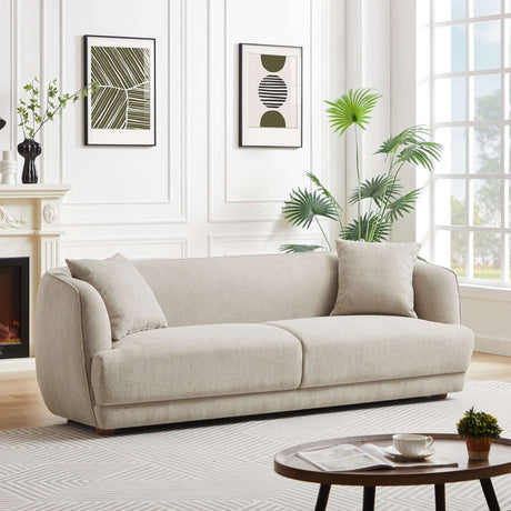 The Larisa Mid-Century Modern Linen Sofa, complemented by cushions, enhances a contemporary living room adorned with two abstract framed artworks and a fireplace with a floral vase. Nearby, a large window illuminates the round coffee table in the foreground stacked with books.