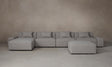 The Breuer Modular Chaise Armless Sectional 114" in Belgian Linen Agate, a spacious and gray-toned modular sectional sofa with multiple cushions, is arranged on a neutral-toned floor against a plain beige wall. The setup includes a matching ottoman positioned slightly to the right.