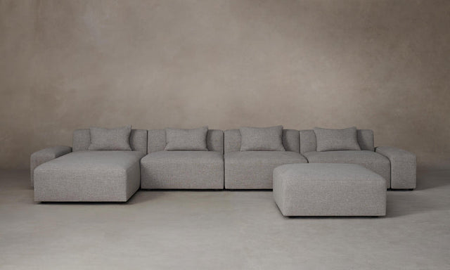 The Breuer Modular Chaise Armless Sectional 114" in Belgian Linen Agate, a spacious and gray-toned modular sectional sofa with multiple cushions, is arranged on a neutral-toned floor against a plain beige wall. The setup includes a matching ottoman positioned slightly to the right.