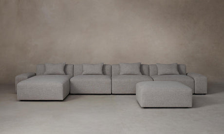The Breuer Modular Chaise Armless Sectional 114" in Belgian Linen Agate, a spacious and gray-toned modular sectional sofa with multiple cushions, is arranged on a neutral-toned floor against a plain beige wall. The setup includes a matching ottoman positioned slightly to the right.