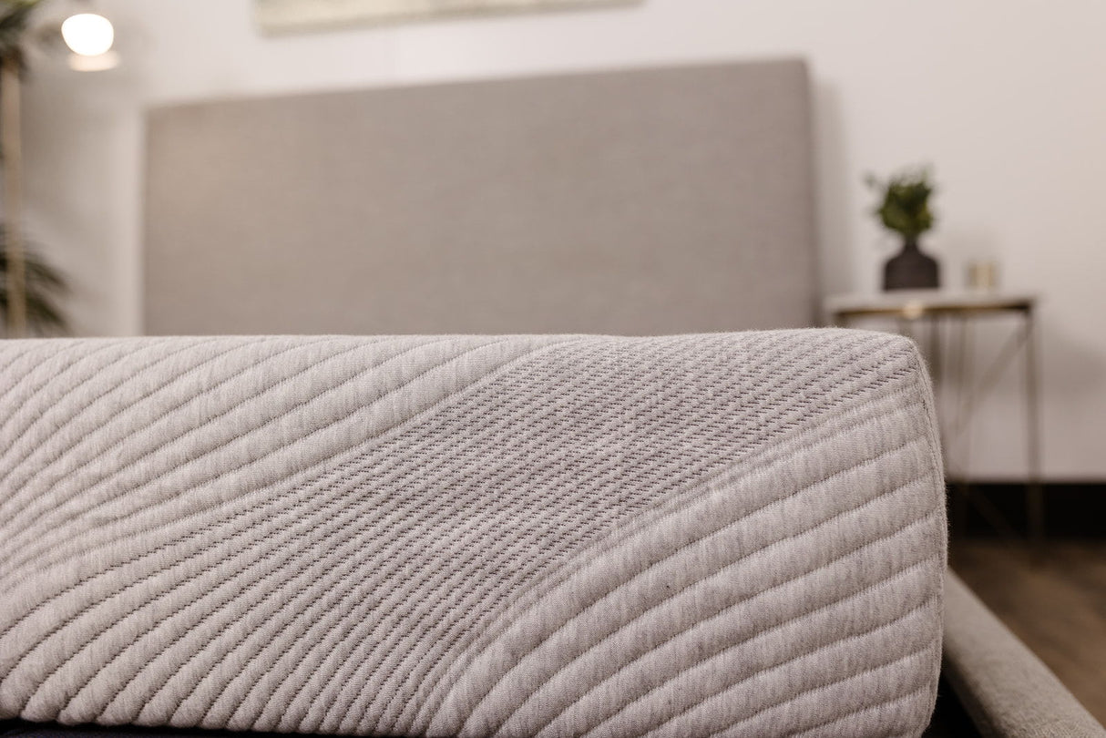 Close-up of the 8" Comfort Series Firm Gel Memory Foam Tight Top Mattress in light gray, featuring a textured, stitched surface, displayed with a matching upholstered headboard and a bedside table with a small plant.