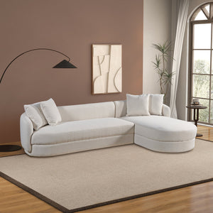 In a modern living room, the Chandler Comfortable Sectional Sofa is set on a beige rug with two cushions. The brown walls feature abstract artwork, accompanied by a floor lamp with a black shade and a tall plant by a large window with white curtains.