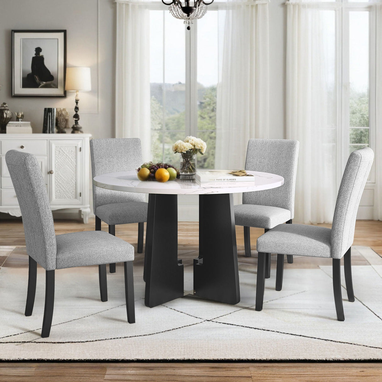 A dining room features a 5 Piece Round Dining Table Set, including a table and four gray upholstered chairs. The space is decorated with flowers and fruit on the table, complemented by a white rug, chandelier, cabinet, and window with sheer curtains.