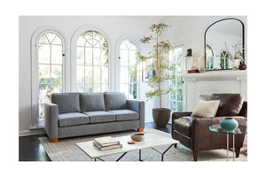 A stylish living room with a Catalina Sofa in gray and a brown leather armchair. A large mirror hangs above a white fireplace adorned with decor. The room features arched windows, a potted plant, and a glass-top coffee table with books and a bowl.