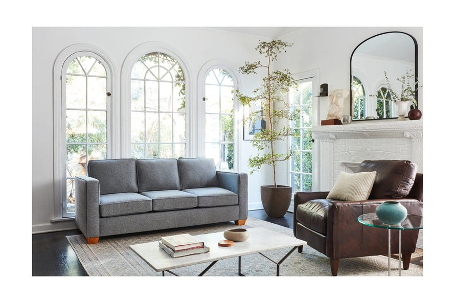 A stylish living room with a Catalina Sofa in gray and a brown leather armchair. A large mirror hangs above a white fireplace adorned with decor. The room features arched windows, a potted plant, and a glass-top coffee table with books and a bowl.