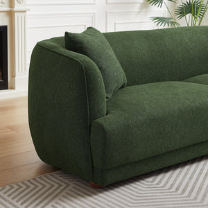 The Larisa Mid-Century Modern Linen Sofa in green, complemented by a matching cushion, sits on a light gray patterned rug. The backdrop includes a white wall with molding and hints of a fireplace and potted plant.